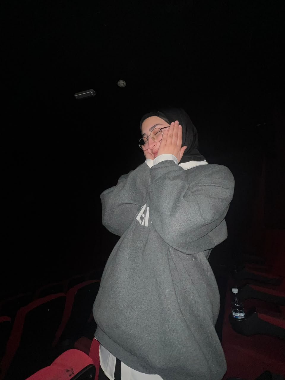 Fatima in an oversized grey sweatshirt in a dark cinema, eyes closed, hands pressed to her cheek.