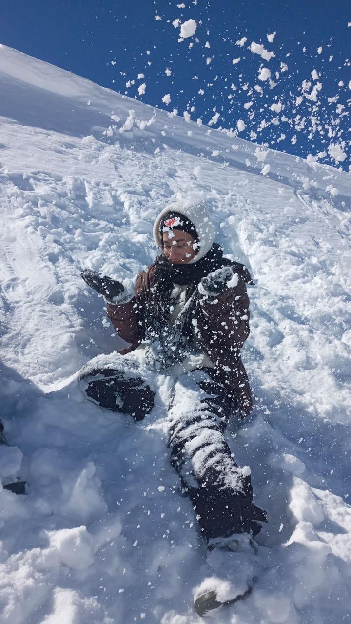 Fatima sitting in deep snow under a bright blue sky, tossing snow into the air.