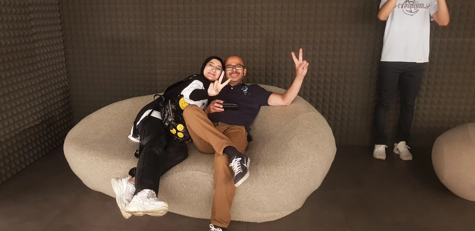 Fatima leaning on a man's shoulder on a big beanbag sofa, both holding up peace signs.