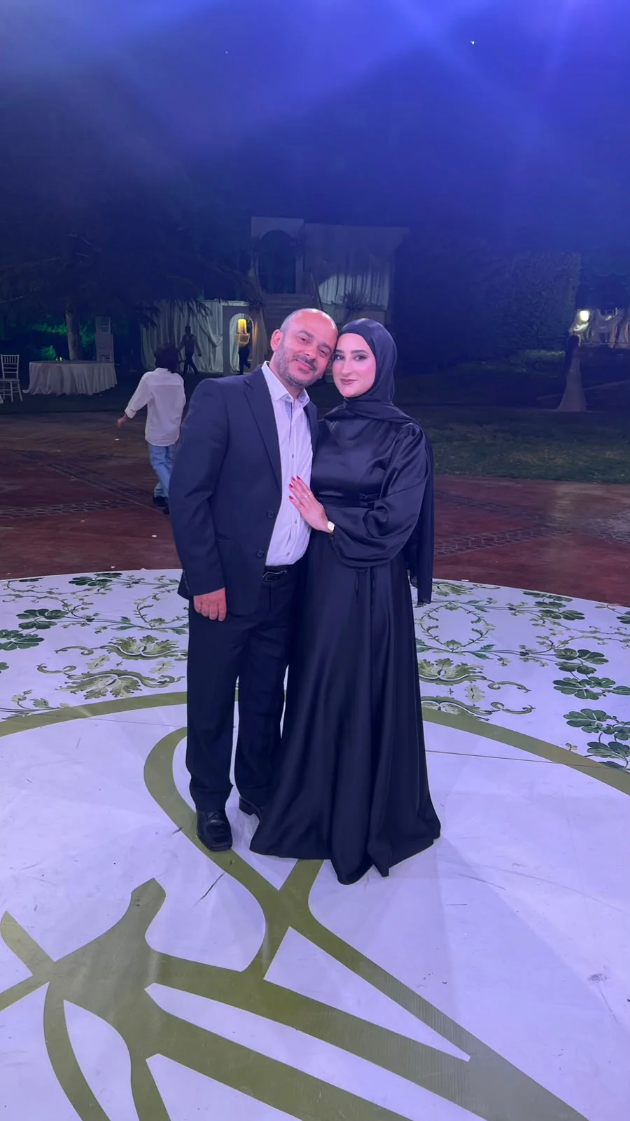 Fatima in a black satin dress standing close beside a man in a suit at an evening celebration.
