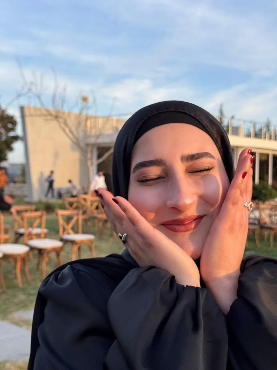 Fatima smiling with her eyes closed, hands resting against her cheeks, in warm evening light.