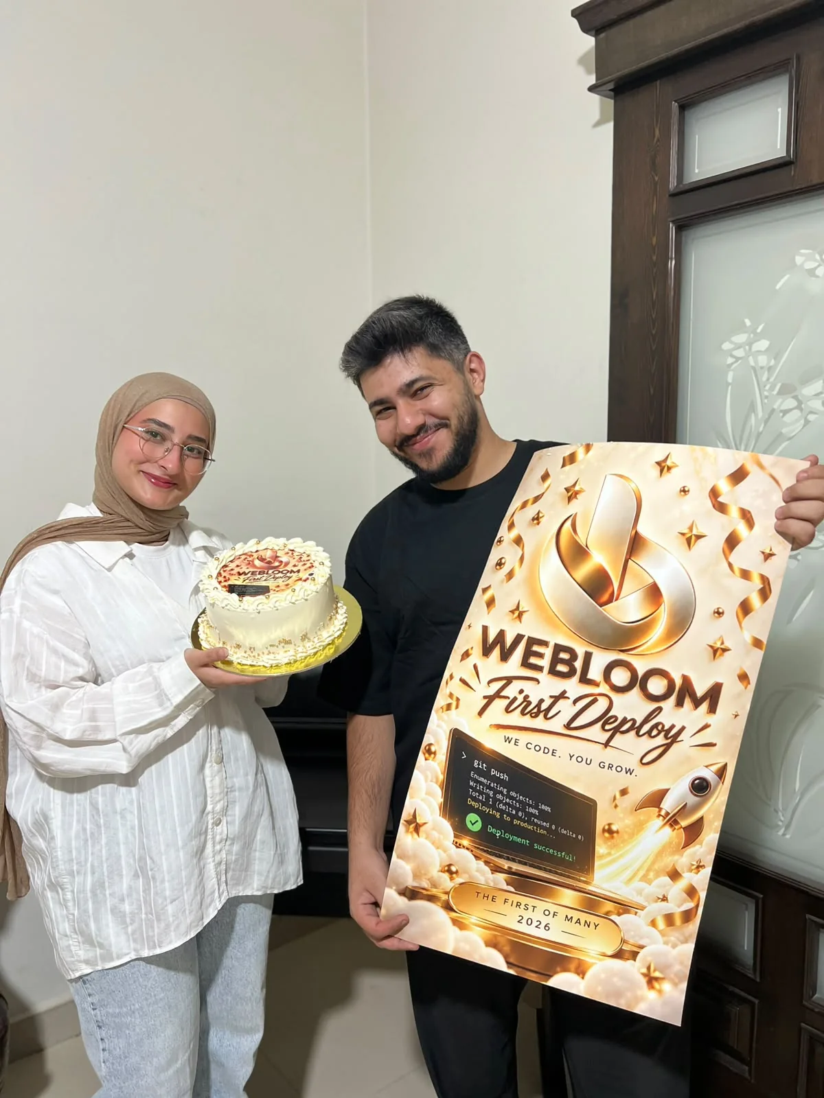 Fatima holding a decorated cake beside a gold poster that reads Webloom First Deploy; both of us smiling.