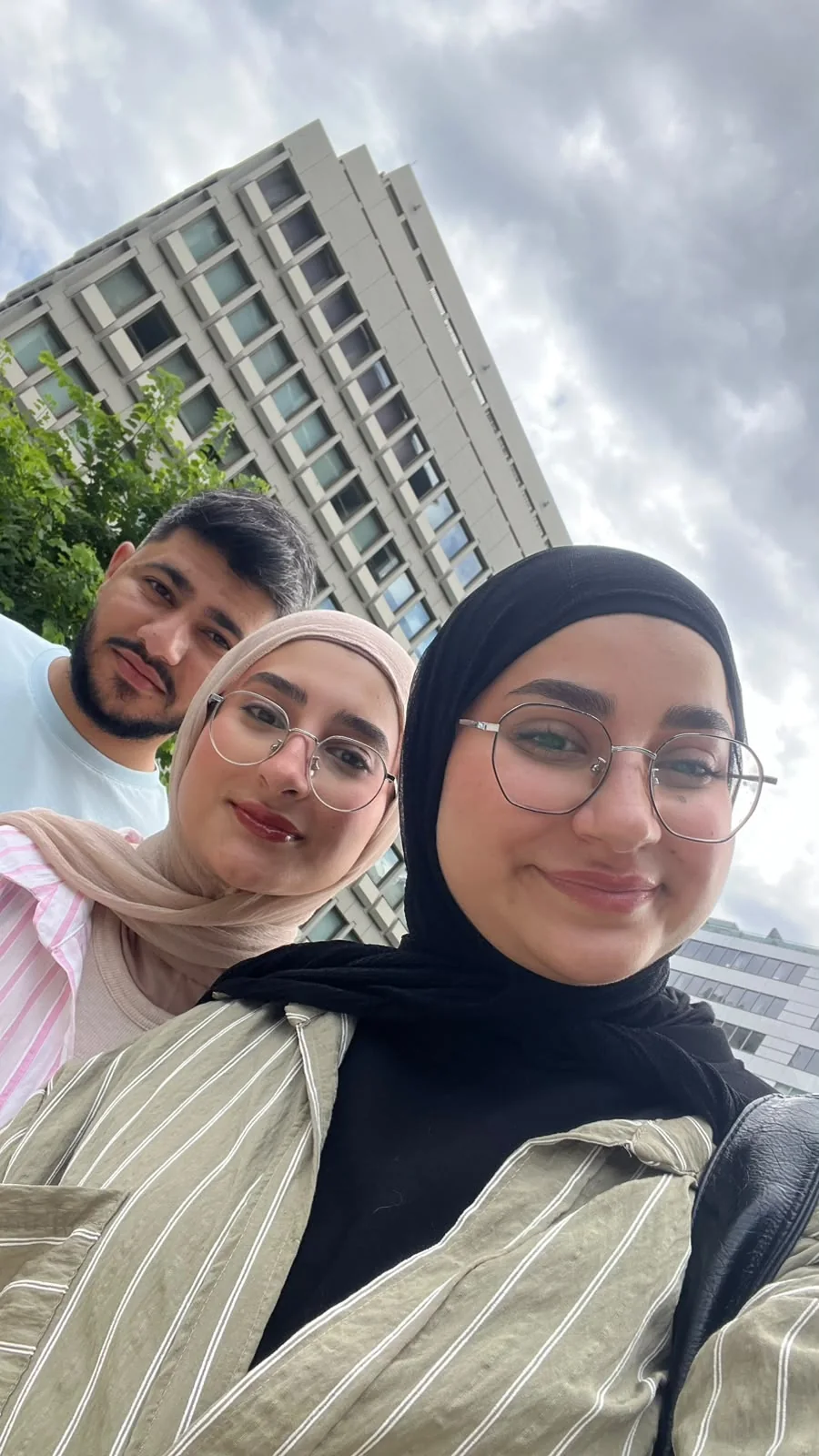 A three-person selfie under a cloudy sky, with a tall building behind.