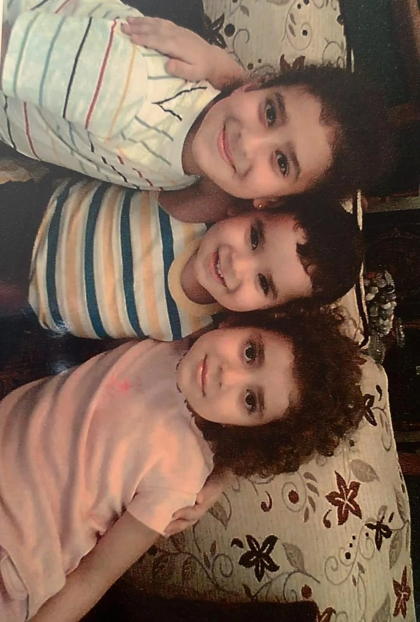 An old printed photograph of three small children smiling, cheek to cheek, on a floral sofa.
