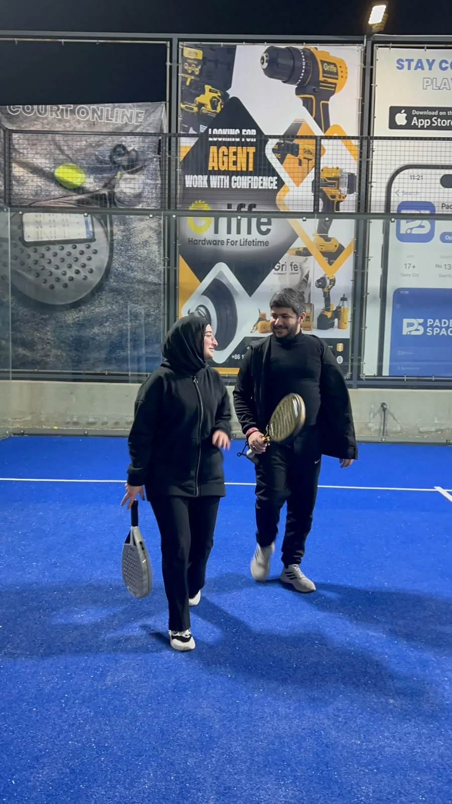 The two of us walking across a blue padel court at night, rackets in hand.