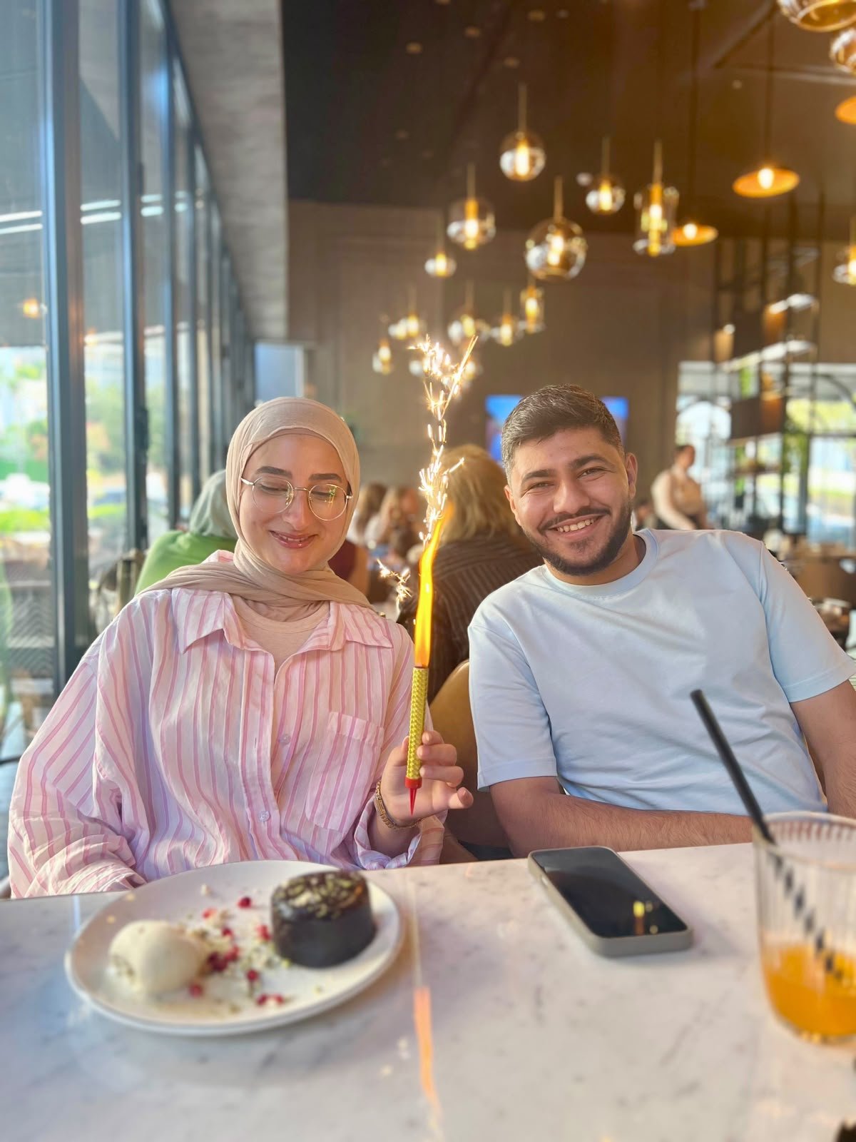 The two of us at a restaurant table; Fatima holds a lit sparkler and smiles down at a dessert.